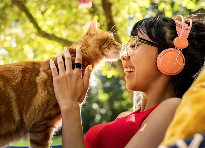 Jeune femme avec casque audio et lunettes souriant à un chat roux en extérieur.