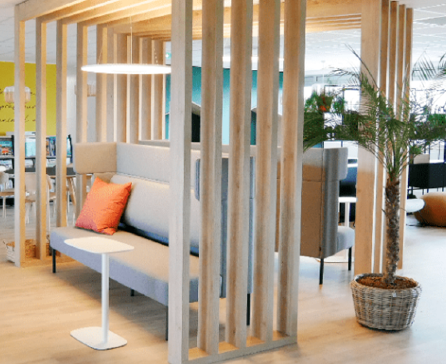 Espace de détente et réunion informelle MSD Coin salon moderne dans un bureau avec banquettes grises, coussin orange, séparations en lattes de bois clair et plante verte en pot.